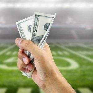 Fist holding dollar bills with 50 yard line on football field in background